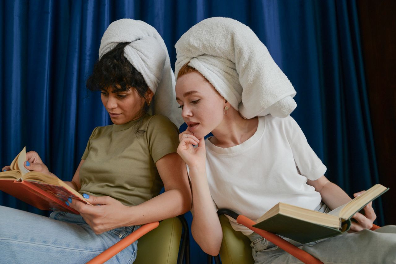 Two women enjoying a calm moment reading with towels on their heads, indoors.